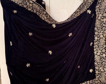 May include: A dark navy blue velvet shawl with detailed gold embroidery. The edges and surface are adorned with floral and paisley patterns. The shawl is draped over a mannequin, highlighting its flowing design and craftsmanship. The overall effect is one of luxury and elegance.