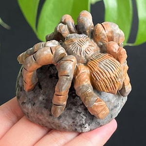 May include: A detailed stone carving of a spider. The sculpture features a gray body and legs with orange and brown accents. The spider is resting on a gray, rounded base. The carving is set against a dark background with green foliage.
