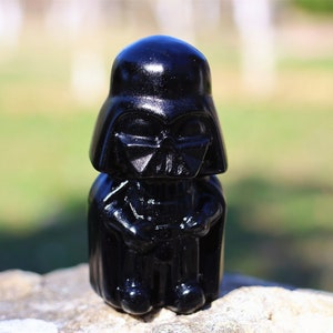 May include: A black plastic figurine of Darth Vader from Star Wars. The figurine is sitting down with its hands clasped in front of it.