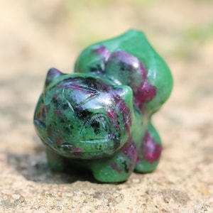 May include: Green and pink carved gemstone figurine of a cartoon character. The figurine is detailed and has a shiny finish.