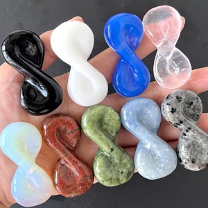May include: A collection of eight infinity symbol-shaped polished stones in various colors, including black, white, blue, clear, green, red, light blue, and gray.
