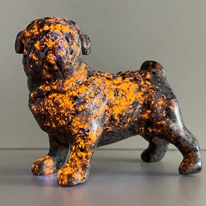 May include: A ceramic pug figurine with a speckled orange and black pattern. The dog is in a standing pose, with its ears perked up and a slight furrow in its brow. The figurine is a decorative item.