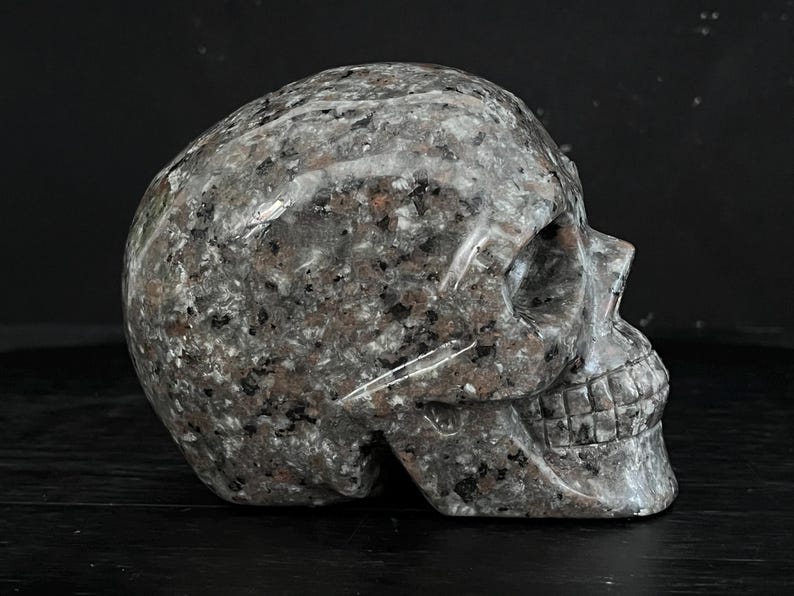 May include: A hand-carved granite skull sculpture. The skull is detailed with a textured surface and features a realistic depiction of the facial structure.