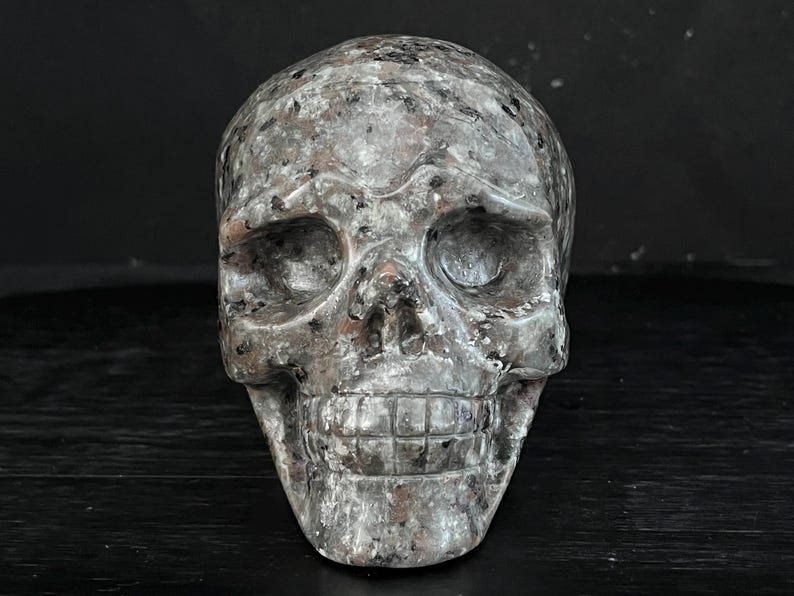 May include: A detailed, hand-carved skull sculpture made from a grey and black speckled stone. The skull has a realistic appearance with defined eye sockets, a prominent nose, and a toothy grin.