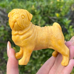 Puede incluir: Figurita de carlino amarillo-naranja detallada. La pequeña estatua de perro está tallada con arrugas y pliegues, y se sostiene en una mano. La figurita es un objeto decorativo.