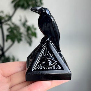 May include: A glossy black raven figurine perched atop a pyramid base. The pyramid features an eye symbol within a triangle. The raven is detailed with a curved beak and textured feathers. The figurine is a decorative item.