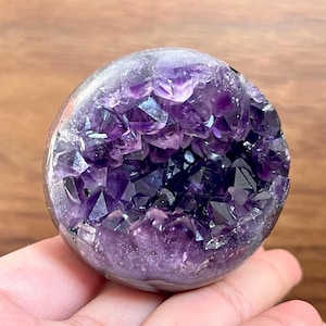 May include: A spherical amethyst geode, showcasing a deep purple crystalline interior. The outer shell is a pale lavender, with the interior crystals catching the light. This natural gemstone is a decorative item.