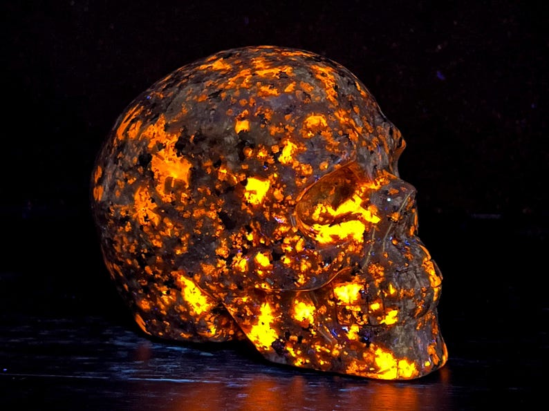 May include: A skull-shaped rock with a dark brown exterior and a bright orange glow emanating from the inside. The rock is covered in small, orange-colored crystals.