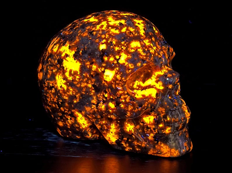 May include: A human skull carved from a dark brown stone with a unique pattern of bright orange and yellow flecks. The skull is illuminated from within, creating a glowing effect.