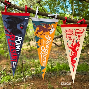 May include: Three felt pennants hang from a tree branch. The pennants are blue, orange, and cream-coloured, each with a unique design and text. The blue pennant features a black bird and the words "Point Pleasant W.V." The orange pennant says "Dine Barrens New Jersey." The cream pennant says "Live Mas."