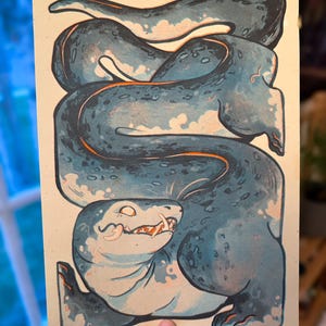 May include: A print featuring a stylized sea creature in blue and white with orange detailing. The artwork depicts a coiled body and a smiling face with teeth. The artist's signature, "KAYCAT," is visible in the bottom right corner.