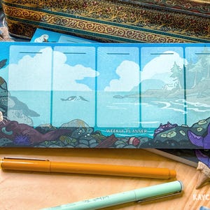 以下が含まれることがあります： 海辺のイラストが描かれた長方形の週間プランナーメモ帳。ラッコ、カモメ、シャチが水中に描かれています。デザインには日々の計画用の5つの縦列があり、中央下部にWEEKLY PLANNERという文字が印刷されています。