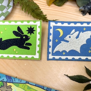Op de afbeelding: Twee geborduurde patches. Eén is groen met een zwart konijn- en sterontwerp. De andere is blauw met een witte vleermuis, maan en sterontwerp. Beide hebben een witte rand.