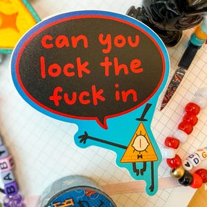 Könnte beinhalten: Ein farbenfroher Aufkleber mit einer Comicfigur und einer Sprechblase. Die Sprechblase ist schwarz mit rotem Text, der lautet: "can you lock the fuck in". Weitere Artikel sind ein Perlenarmband und ein Stift.