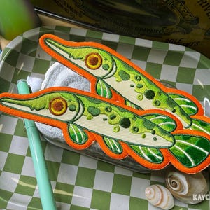 Puede incluir: Dos parches bordados de pez con hocico largo, vientre color crema y escamas verdes moteadas. Cada parche está delineado con un borde naranja vibrante. Los peces tienen ojos circulares marrones y costuras detalladas en las aletas, presentados en par.