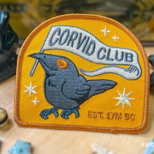 May include: A semi-circular embroidered patch with a mustard yellow background. It features a dark gray crow holding a white banner that reads CORVID CLUB in dark gray text. The patch includes white star accents and the text EST. 17M BC.