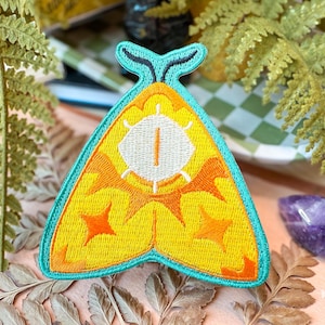 Puede incluir: Parche de polilla amarilla bordada con detalles de estrella naranja y un diseño de ojo blanco. El parche tiene un borde turquesa y una parte superior en forma de antena turquesa. El parche está sobre una superficie con hojas de helecho y un cristal púrpura.