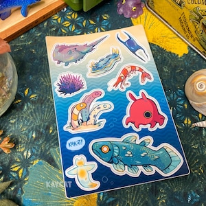 May include: A sheet of stickers featuring various sea creatures, including a seahorse, a starfish, a jellyfish, a sea slug, a shrimp, a sea anemone, and a fish. The stickers are on a blue background with white outlines. The text "Kayak!" is written on one of the stickers.