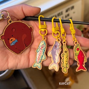 May include: Wooden keychains featuring a bear with a fish and four colorful fish designs. The bear keychain is round and red. The fish keychains are various colors, including blue, purple, and red, with yellow hardware.