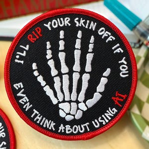 Puede incluir: Un parche circular negro con borde rojo, con un diseño de mano de esqueleto bordada en blanco y texto. El texto dice: "I'll rip your skin off if you even think about using AI."