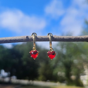 May include: A pair of gold hoop earrings with red heart-shaped charms hanging from them. The earrings are photographed against a blue sky with white clouds.