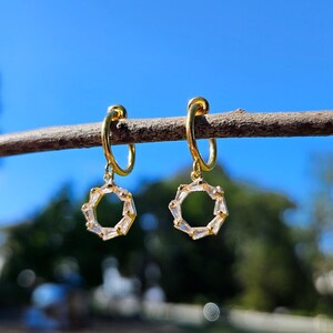 May include: A pair of gold hoop earrings with a dangling circle design. The circle is made of small, clear cubic zirconia stones.