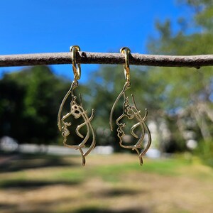 May include: Gold hoop earrings with a delicate, abstract face design. The earrings are hanging from a branch against a blue sky and green foliage background.