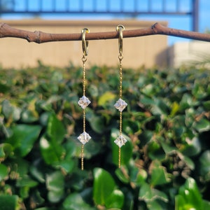 May include: A pair of gold hoop earrings with a dangling chain and three square-shaped clear crystals.