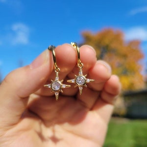 May include: A pair of gold hoop earrings with a star-shaped charm featuring a clear crystal in the center. The star has smaller crystals around the edges.