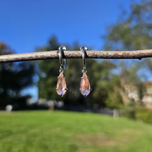 May include: A pair of silver hoop earrings with teardrop-shaped, peach-colored gemstones. The earrings are hanging from a brown branch against a backdrop of a bright blue sky and green grass.