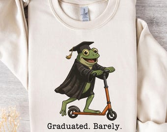 May include: Cream-colored sweatshirt featuring a cartoon frog wearing a graduation cap and gown, riding an orange scooter. The text "Graduated. Barely." is printed below the frog. The sweatshirt has long sleeves and a crew neck.