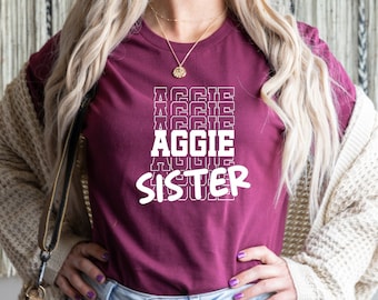 May include: A maroon t-shirt with white text that reads "Aggie Sister" in a stacked, repeating pattern.