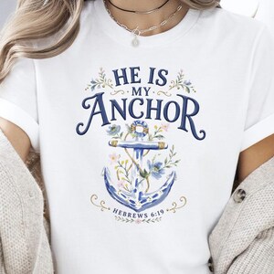 May include: A white short-sleeved t-shirt featuring a blue graphic of an anchor entwined with colorful floral illustrations. The text reads HE IS MY ANCHOR and HEBREWS 6:19 in a dark blue font, centered on the chest of the garment.