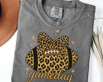 May include: Gray Comfort Colors t-shirt with a leopard print football graphic and the word "gameday". The football has a bow and star accents. A silver necklace with a pendant is also visible.