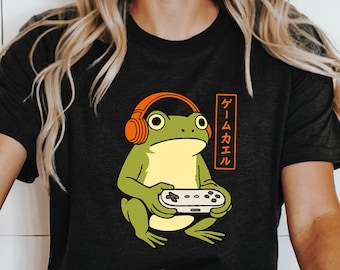 May include: Dark gray t-shirt featuring a cartoon frog wearing orange headphones and holding a video game controller. The frog is green and yellow. Japanese text in an orange rectangle is on the right side of the frog.