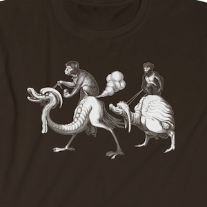 Puede incluir: Una camiseta marrón con una ilustración en blanco y negro de tres monos montando una criatura mítica.