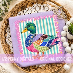 Könnte beinhalten: Ein lilafarbenes T-Shirt mit einer farbenfrohen Entenillustration. Die Ente hat einen grünen Kopf, einen blau geblümten Körper und Flickenflügel. Der Text "1/2 Penny Digitals Original Designer" ist unten aufgedruckt. Das T-Shirt liegt auf einem Weidenkorb.