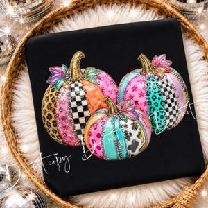 May include: A black t-shirt featuring a graphic of three pumpkins decorated with colorful patterns including leopard print, black and white checkers, pink stars, polka dots, and cow print, accented with gold stems and vibrant floral details.