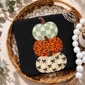 Op de afbeelding: Een zwart sweatshirt met een stapel van drie pompoenen. De bovenste pompoen heeft een saliegroen en wit ruitpatroon, de middelste is oranje met een zwarte luipaardprint en de onderste is crème met een patroon van zwarte strikjes.