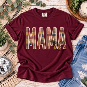 May include: A maroon short-sleeve t-shirt featuring the word MAMA printed in large, block letters. The letters have a multi-colored plaid pattern in shades of red, yellow, blue, and green. The shirt has a crew neckline and a Comfort Colors label.