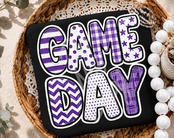 May include: A black t-shirt featuring the text GAME DAY in large, bold, white-outlined letters. Each letter displays a different purple and white pattern, including horizontal stripes, polka dots, plaid, stars, and chevron zig-zag designs.