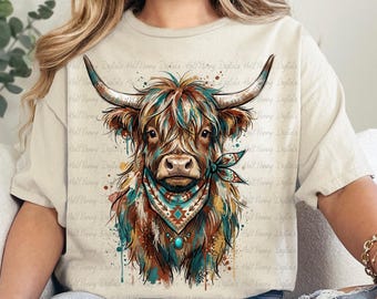 May include: A beige t-shirt featuring a watercolor illustration of a Highland cow. The cow has long horns, a colorful bandana, and a turquoise pendant. The design includes splashes of teal, brown, and orange. The text "Half Finny Digitals" is visible.