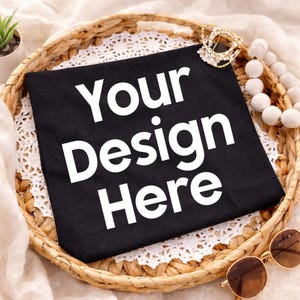 Könnte beinhalten: Schwarzes T-Shirt mit dem weißen Text "Your Design Here", ordentlich in einem geflochtenen Korb gefaltet. Der Korb ist mit einer weißen Spitzendecke ausgelegt. Zubehör: goldene Perlenkette und Sonnenbrille.