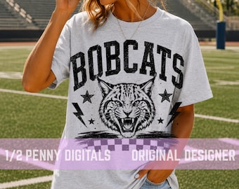 May include: A light gray short-sleeve t-shirt featuring a black graphic of a snarling bobcat face. The text BOBCATS is printed in a bold, distressed font above the animal. The design includes lightning bolts, stars, and a checkered pattern.