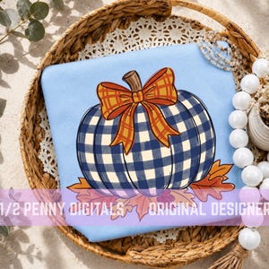 Op de afbeelding: Een lichtblauw sweatshirt met een afbeelding van een pompoen met een marineblauw en wit ruitpatroon, bekroond met een oranje en gele geruite strik, rustend op oranje herfstbladeren. De tekst 1/2 PENNY DIGITALS ORIGINAL DESIGNER is zichtbaar.