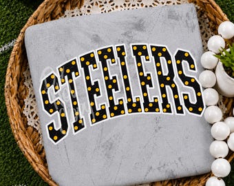 May include: A light gray sweatshirt featuring the word STEELERS in a bold, arched font. The letters are black with a white outline and are filled with a pattern of small yellow polka dots. The garment is displayed flat within a woven basket.