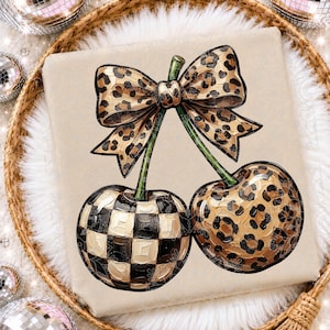 May include: A beige t-shirt featuring a graphic of two cherries connected by a leopard print bow. One cherry has a black and white checkered pattern, while the other cherry features a brown and black leopard print design with green stems.