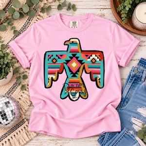 Puede incluir: Una camiseta de manga corta de color rosa claro con un gran gráfico de un águila con las alas extendidas. El águila está rellena con un patrón geométrico colorido en tonos turquesa, naranja, rosa y negro. La camiseta tiene cuello redondo.