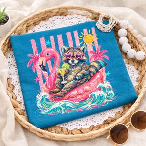 Op de afbeelding: Teal t-shirt met een wasbeer met zonnebril op een roze flamingo-vlot, met een cocktail. Het ontwerp bevat een zon, palmbomen en golven. Het shirt ligt op een vlechtwerk dienblad.