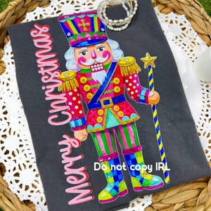 Puede incluir: Una camiseta gris oscuro con un gráfico de cascanueces multicolor vibrante, que sostiene un bastón con una estrella dorada y el texto Merry Christmas en letra rosa. El diseño incluye patrones brillantes en rosa, verde, azul y amarillo.